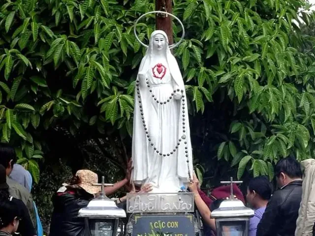 A white robed statue with a prayer rosary, carried on a platform by a crowd