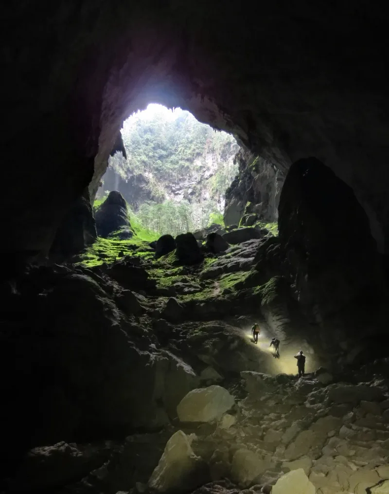 A vast chamber inside Son Doong cave, daylight falling through a ceiling opening onto tiny figures below