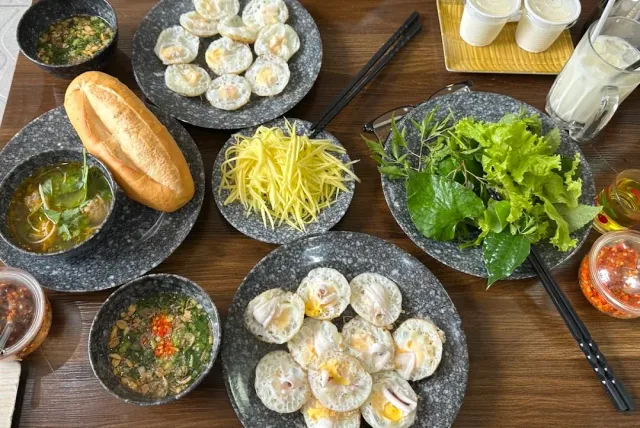 A table with plates of small pancakes, a baguette, shredded green mango and bowls of broth