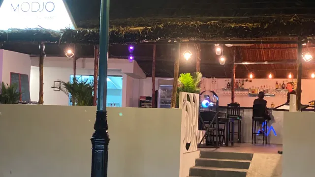 A thatched roof bar at night, purple lighting and bottles on the shelf behind the counter