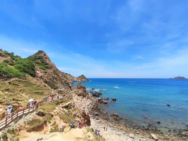 A rocky coastline with a wooden railed path along the cliff, visitors and deep blue sea