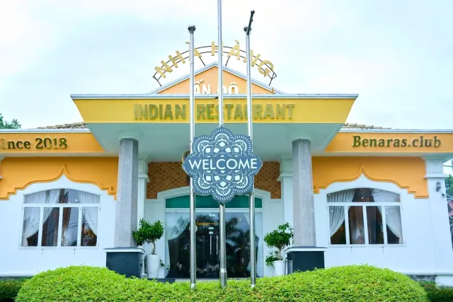 A yellow and white building with a golden sign, a round medallion between flagpoles and trimmed hedges