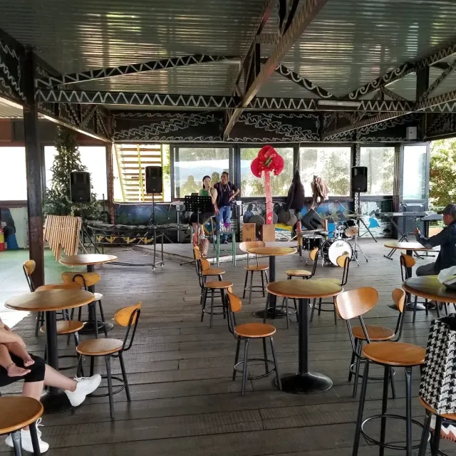 A covered wooden deck with high round tables, a small stage with drums and glass walls