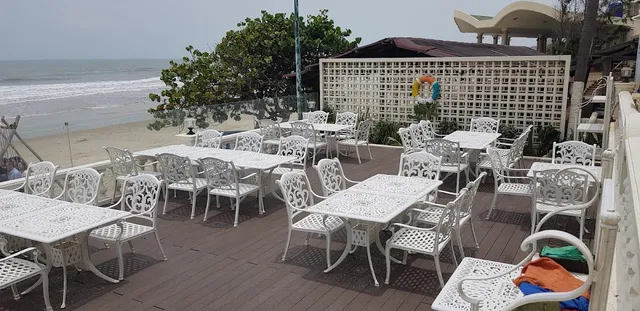 A wooden terrace with ornate white tables and chairs, facing a beach and waves