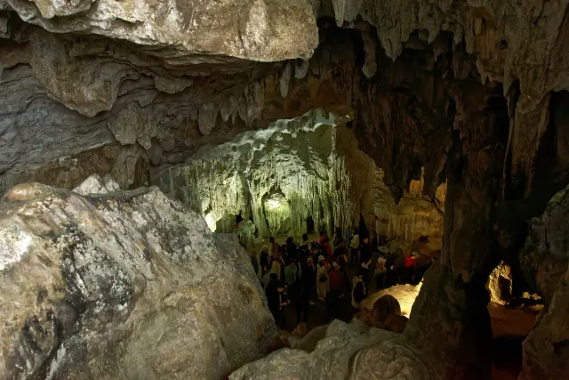 A lit cave chamber with stalactites and stalagmites and visitors on a walkway