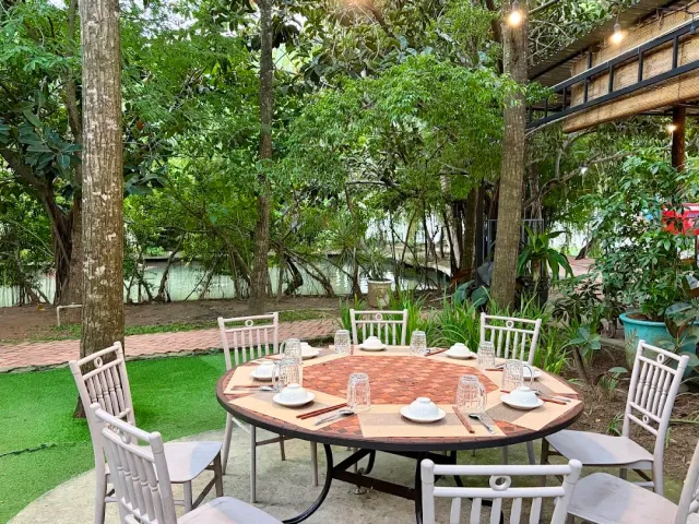 A set round table with white rattan chairs among trees, water in the background