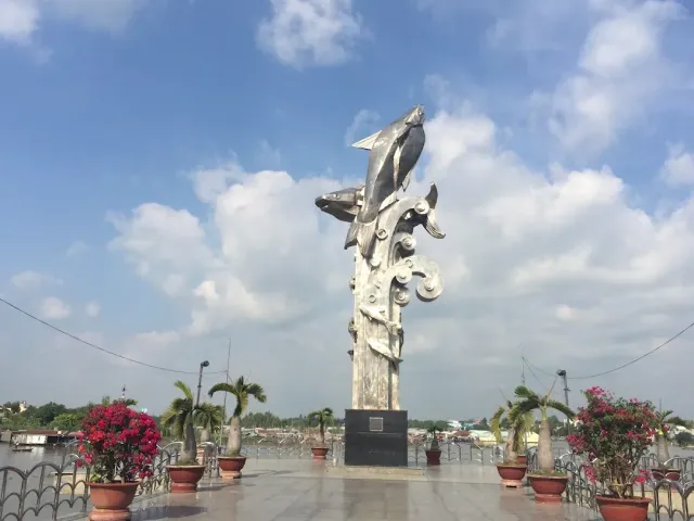 A silver metal monument shaped like two fish on a pillar, on a paved plaza facing a river