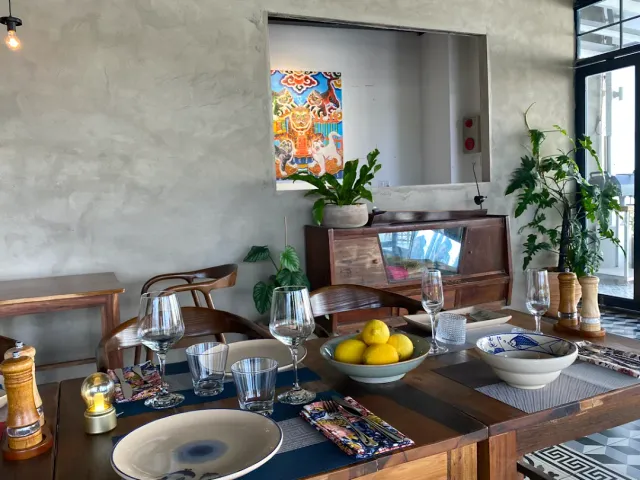 A wooden table set with glasses and a bowl of lemons, a concrete wall with a colorful print and plants
