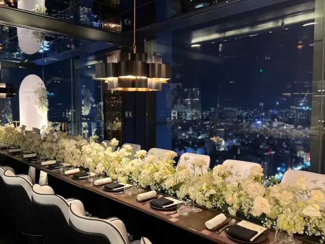A long table set with white flowers beside a glass wall with a night city view