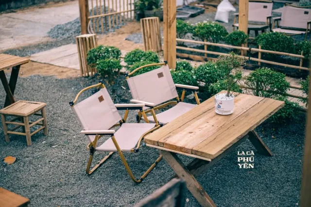 A gravel yard with light folding canvas chairs, a wooden table and trimmed shrubs around