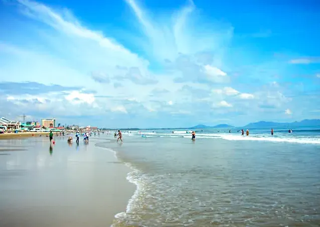 A strip of wet sand with bathers in the shallows and low buildings in the background