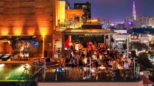 A rooftop at night with a pool, tables crowded with guests and a tall tower on the skyline