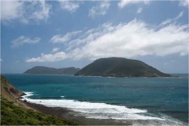 A green landmass across a rough blue sea, white waves breaking on a pebble shore