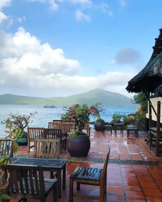 A tiled terrace with wooden chairs and potted frangipani trees, the sea and a green mountain opposite