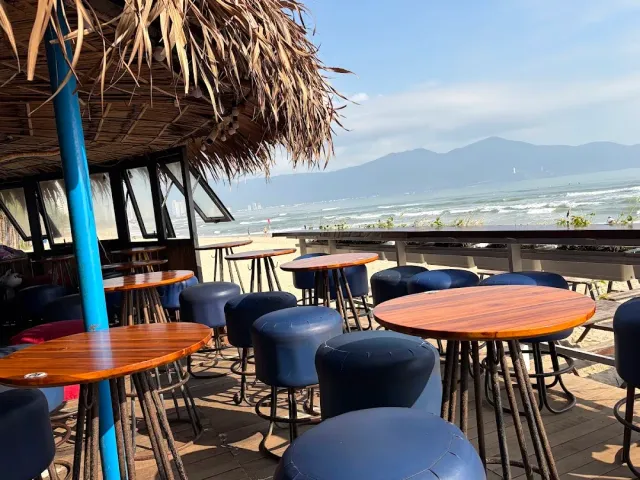 A wooden deck with round tables and blue stools under a thatched roof, the sea and mountain behind