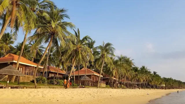 Palms leaning over golden sand, tile roofed bungalows and people walking along the shore