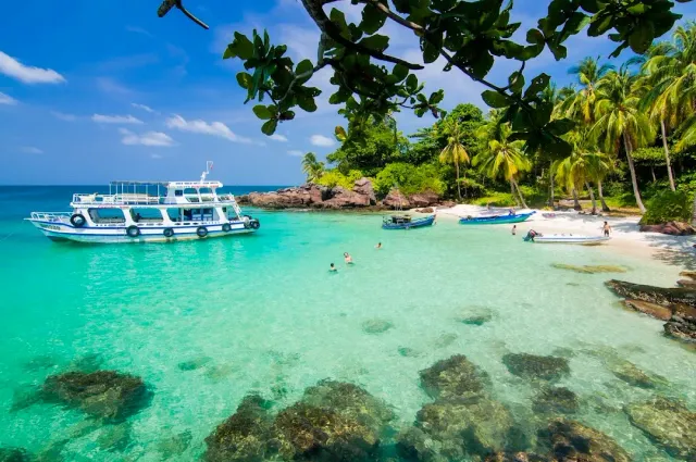 A clear turquoise bay with rocks under the water, a white boat and a palm beach