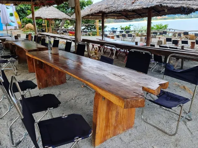 A thick wooden table and canvas chairs on the sand, thatched umbrellas and a greenish sea behind