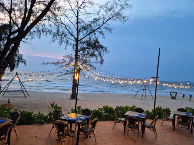 A terrace at dusk facing the beach, strings of bulbs between poles and rattan tables