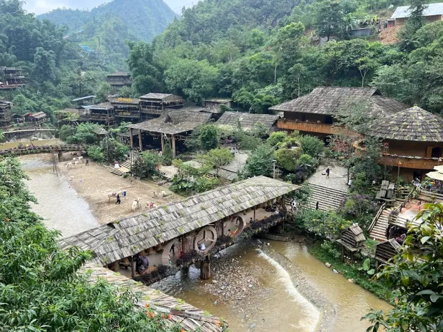 A view from above of wooden houses in a valley and a long covered bridge over a muddy stream