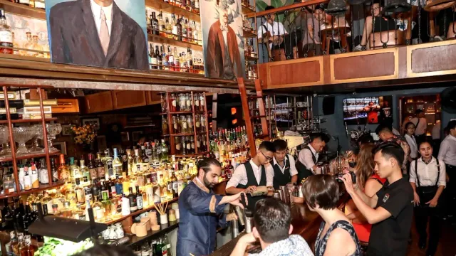 Bartenders in suits working behind a counter packed with bottles, beneath painted portraits