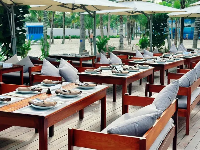 A wooden deck with parasols, tables set with folded napkins and benches facing palms and sand
