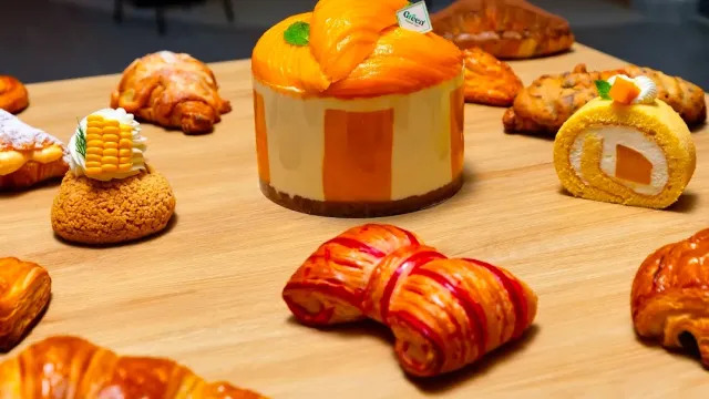 An orange mousse cake at the center of a wooden surface, surrounded by croissants, pastries and a roll slice