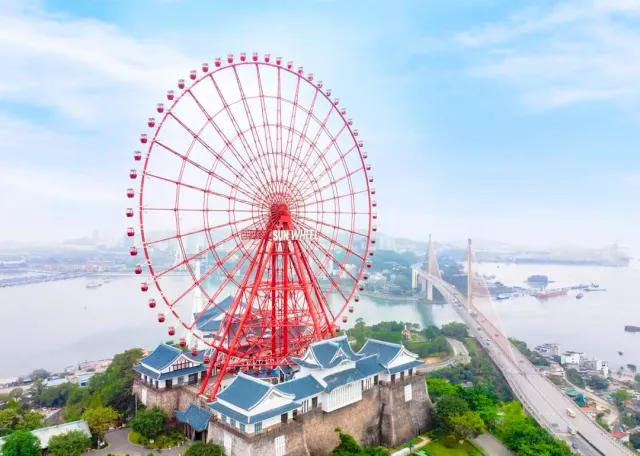 A giant red wheel on a hill above blue roofed buildings, and a misty bay behind