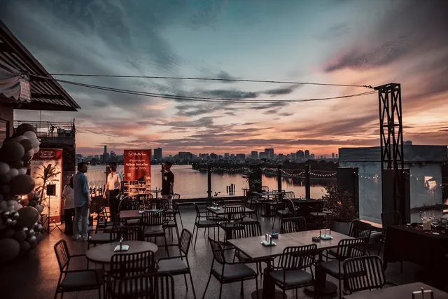 A rooftop terrace with tables facing a lake and a skyline at a pink sunset