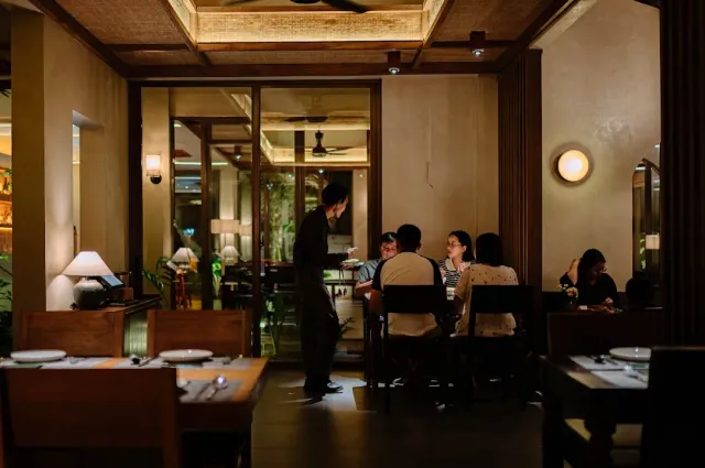 A dim hall with a coffered wooden ceiling, a waiter serving a table of diners and warm lamps