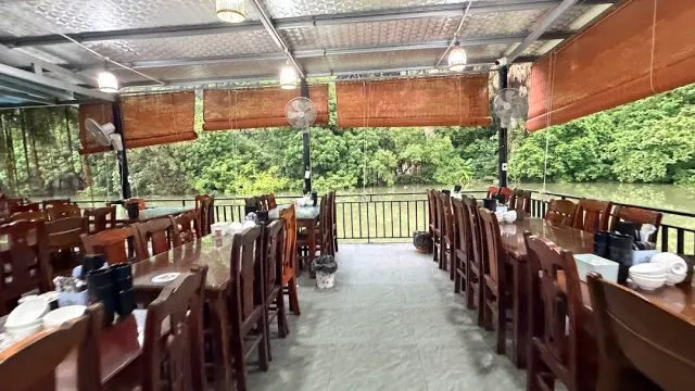 An open hall with two rows of wooden tables, bamboo blinds and a green view to the river