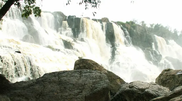 A wide waterfall over semicircular rock steps with boulders in the foreground