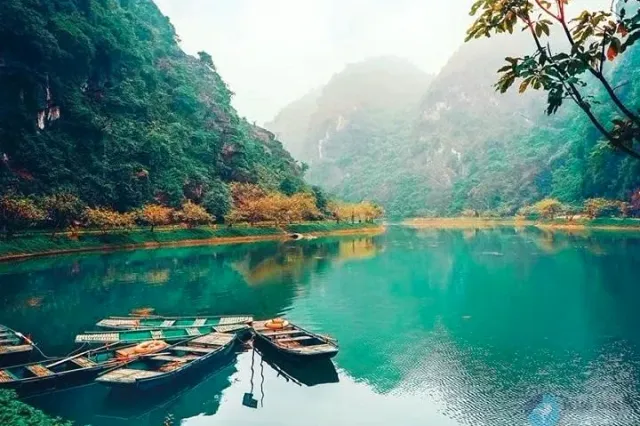 An emerald pond with moored wooden boats, limestone cliffs and trees reflected in the water