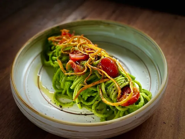 A bowl of pasta in green sauce with cherry tomato halves and crisp fried onion rings