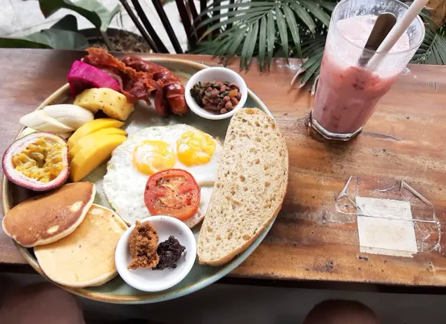 A breakfast plate with fried eggs, bacon, pancakes and tropical fruit beside a pink shake