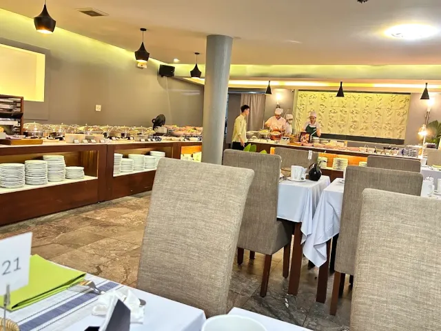 A buffet counter with serving trays and stacks of plates, upholstered chairs and tables in white cloths