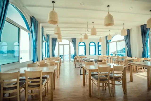 A bright hall with blue curtains, arched windows onto the sea and woven hanging lamps