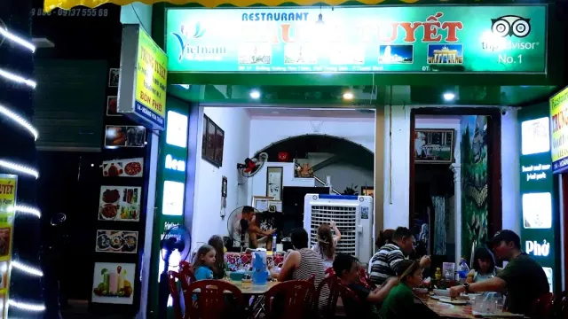 A restaurant front in the evening with a green lit sign, diners at red plastic tables