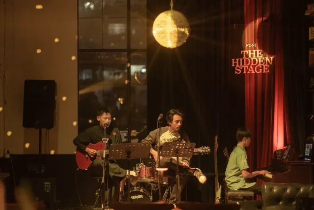 Three musicians with guitars and keyboard on a small stage, a disco ball and a red curtain