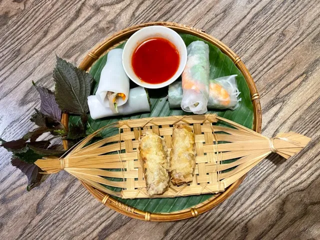 A round wicker tray on a banana leaf with fresh rice rolls, fried rolls and a small bowl of red sauce