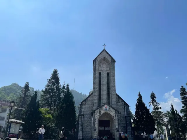 A grey building with a pointed bell tower topped by a cross and conifers on both sides