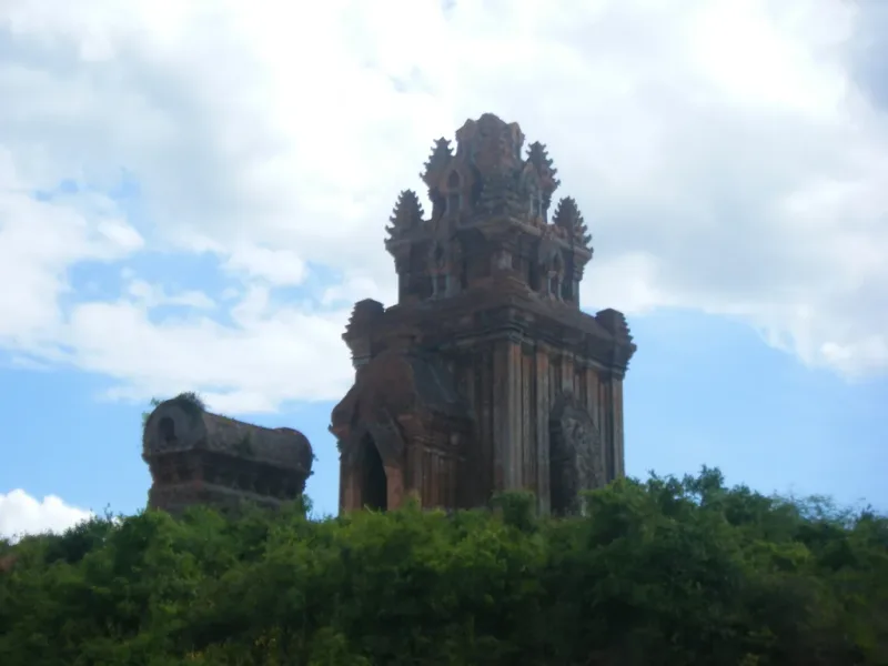The main red brick Cham tower with a lower saddle roofed gate tower beside it, on a green hilltop under cloudy sky