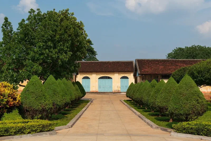 A path between clipped shrubs to the yellow prison building with turquoise doors, at the former Buon Ma Thuot prison