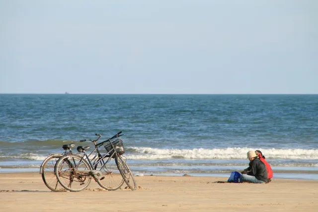 Bicycles on the sand beside people sitting, grey sea and overcast sky