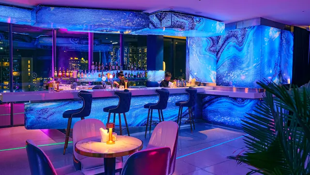 A blue lit bar counter with stools and pink armchairs by the windows