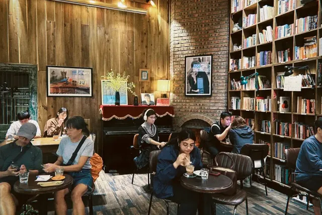 A wood paneled room with bookshelves to the ceiling, an upright piano and guests around tables