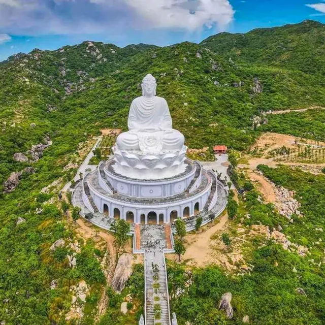 A view from above of a huge white Buddha on a round base atop a green hill