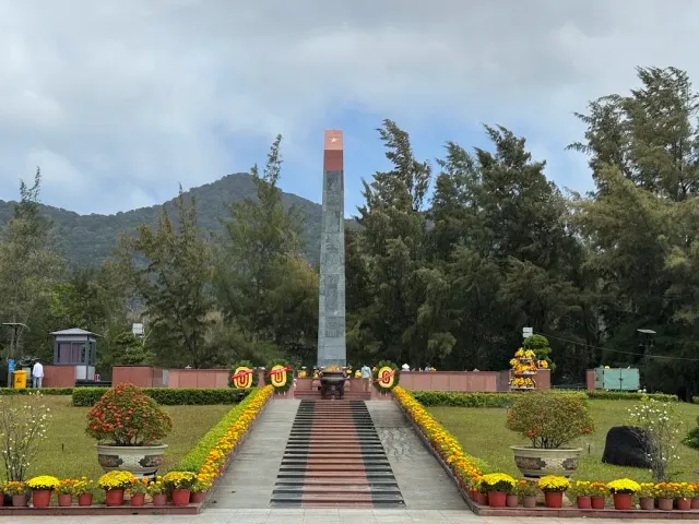 A tall stone obelisk at the top of a stairway, between rows of yellow chrysanthemum pots and green trees