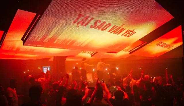 A club with red screens on the ceiling and a dense crowd raising hands facing the stage