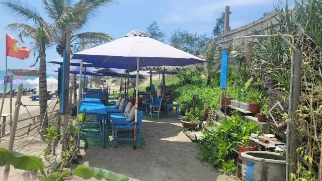 A large umbrella over blue wooden tables and chairs on the sand, palms in the background